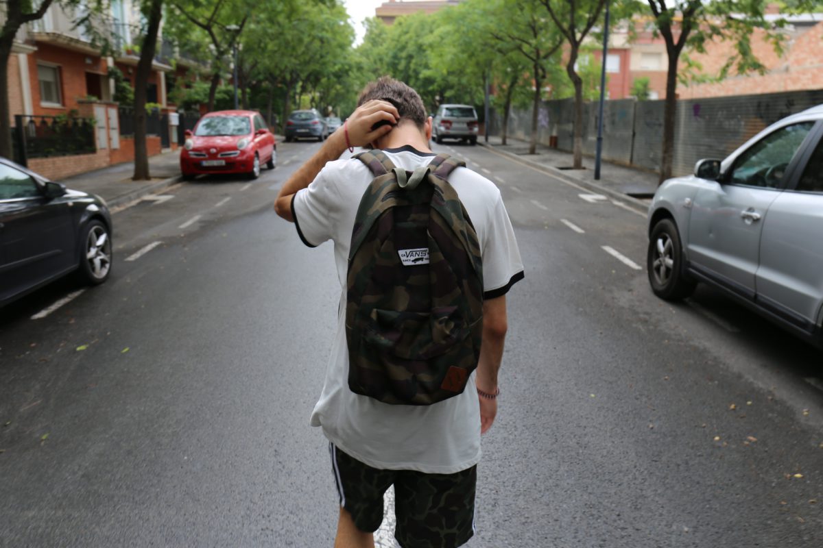 Shot from the back of a boy in t-shirt and shorts, carrying a backpack and rubbing at his head