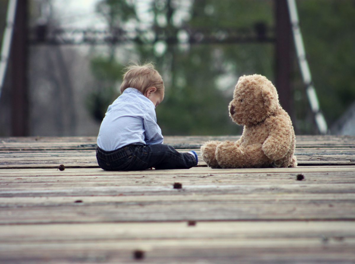 Image of a young toddler sitting with a stuffed teddy bear on a wood surface
